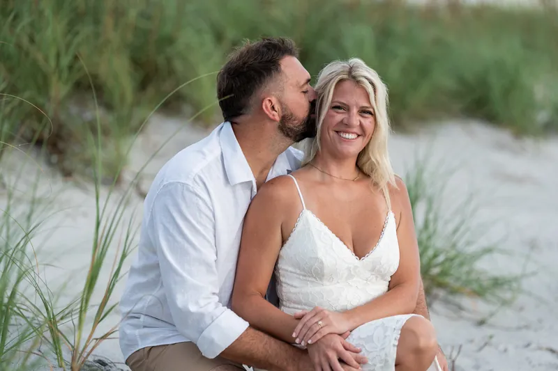 Brandon kisses Jarah's cheek as they sit together in the sand dunes with beach grass behind them.