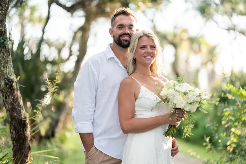 Jarah and Brandon smile together under sprawling oaks, Jarah holding a white bouquet at Mussallem Beachfront Park.