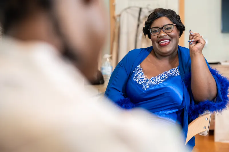 Jazzalynn smiles at her reflection in a mirror while adjusting her hair, wearing glasses and a blue wrap.