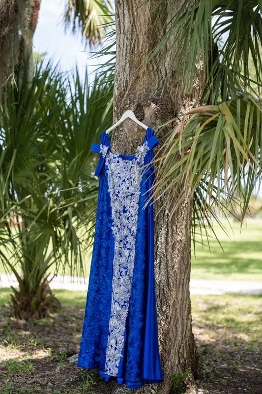 Person in a blue and white patterned dress stands with arms wrapped around a large tree trunk surrounded by palm fronds.