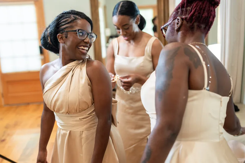 Three people in cream and beige bridesmaid dresses chat indoors, with bright windows behind them.