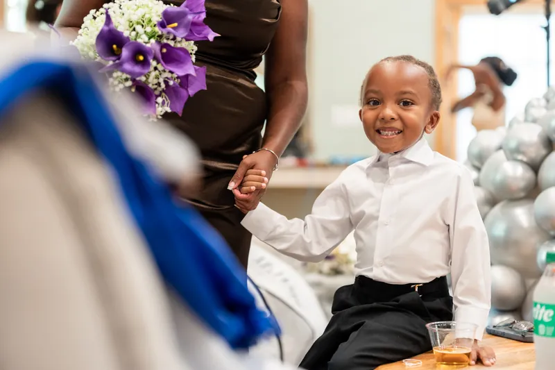 A young child in a white dress shirt and black pants holds hands with someone in a brown dress holding a purple and white