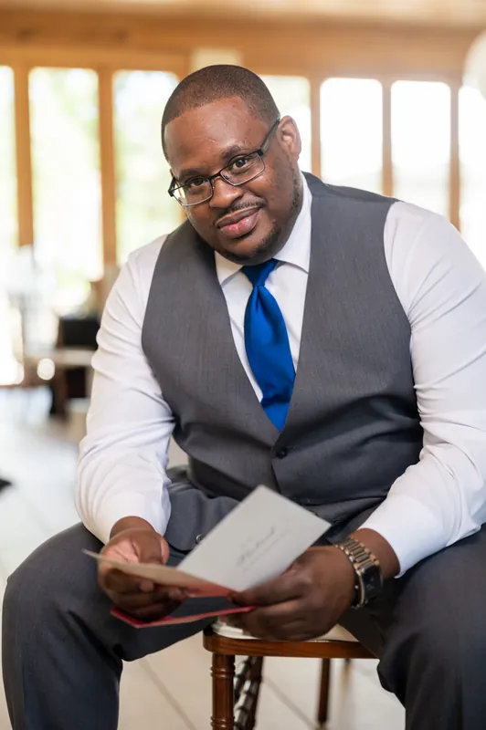 Hakeem sits on a wooden chair in a gray vest and blue tie, holding a program and smiling at the camera.