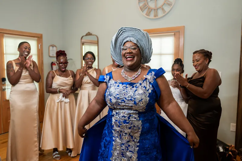 A woman in a vibrant blue and white patterned dress with a coordinating head wrap stands with arms outstretched, surrounded