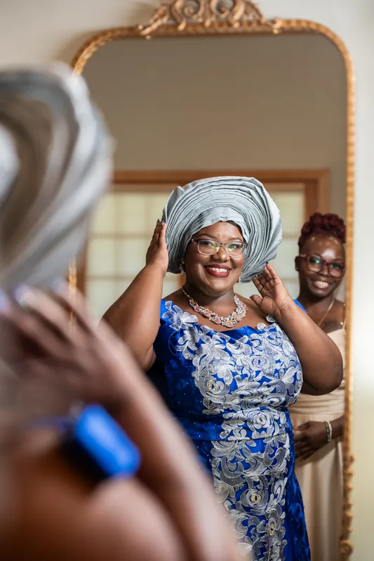 A woman in a blue and white patterned dress adjusts her matching head wrap while looking in a mirror, with two people visible