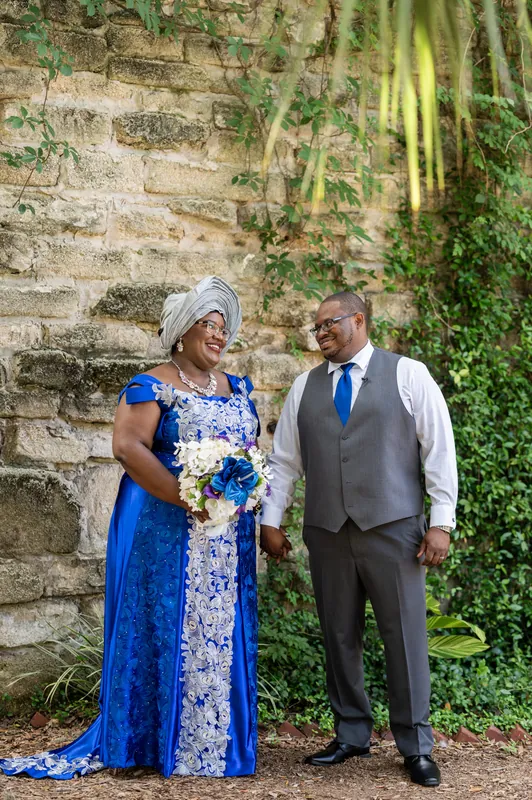 Jazzalynn in a blue patterned dress and Hakeem in a gray vest stand together against a moss-covered stone wall, holding