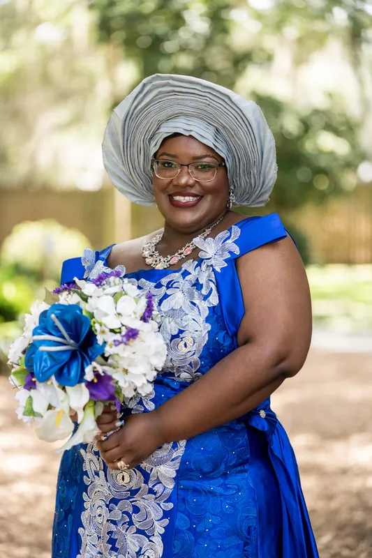 Jazzalynn wears a bright blue dress, gray head wrap, and glasses while holding a white and blue bouquet and smiling