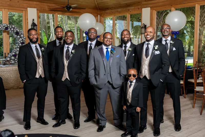 Hakeem stands center with nine groomsmen in dark suits and ties, all posed together inside a wooden pavilion.