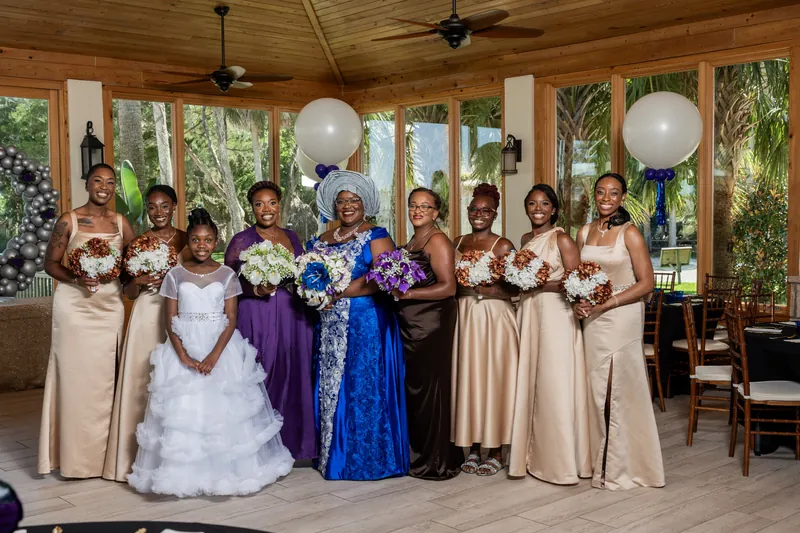 A wedding party of fourteen stands in a line inside the pavilion, wearing neutral and jewel-toned formal attire with white