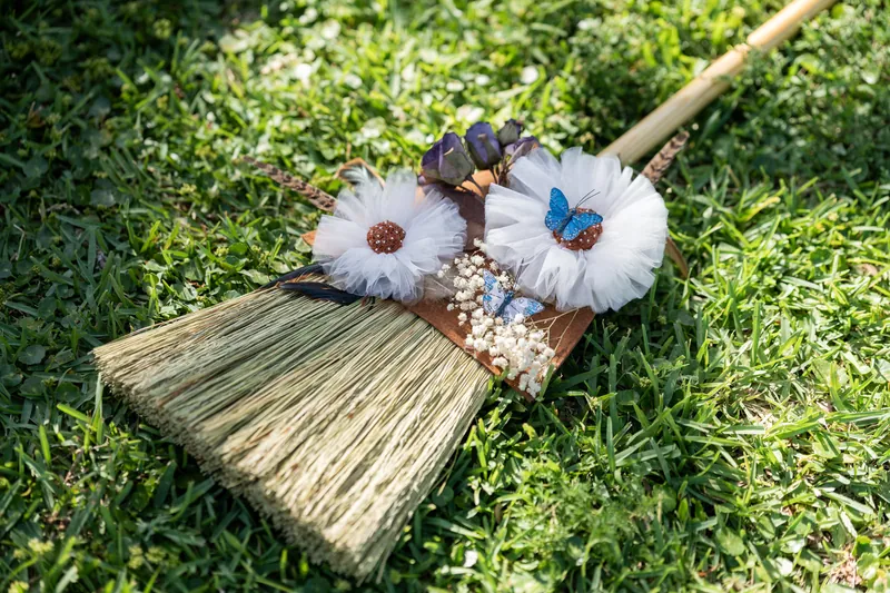 White flowers with blue butterflies rest on a small woven broom beside green grass and a wooden pole.