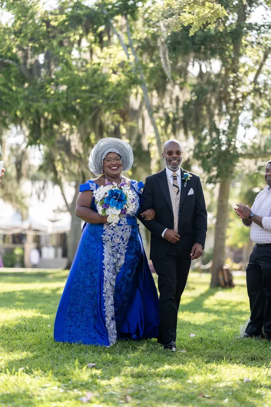 A woman in a blue dress and a man in a dark suit walk together on the grass beneath Spanish moss-draped trees.