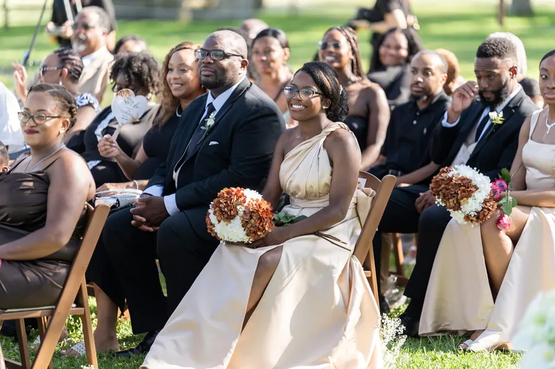 Attendees in formal attire hold bouquets and programs while seated on chairs during the outdoor ceremony.
