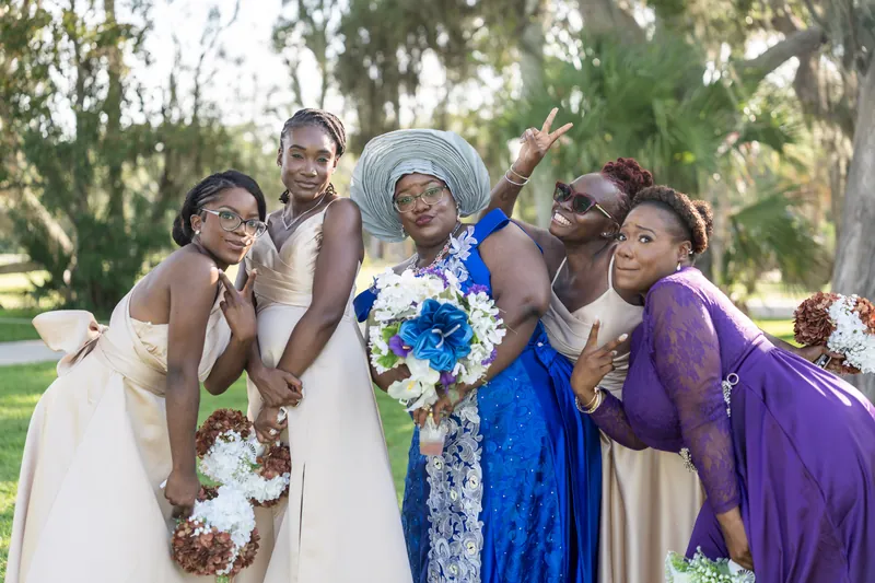 Four women in various colored dresses laugh together, each holding bouquets of white and blue flowers outdoors.