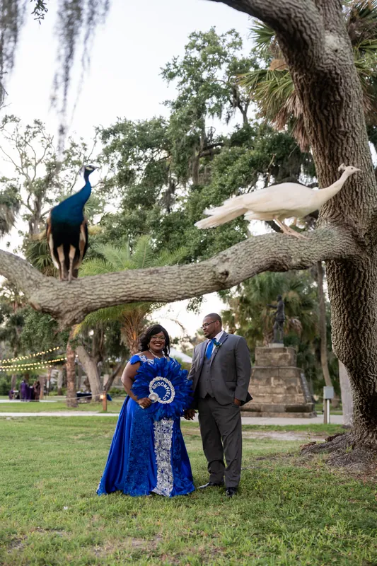 Jazzalynn and Hakeem stand together beneath a sprawling oak tree with hanging moss, a peacock perched on a branch above them.