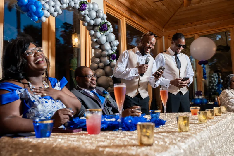 Guests laugh and chat at the reception table, decorated with blue balloons and glassware, as speakers stand