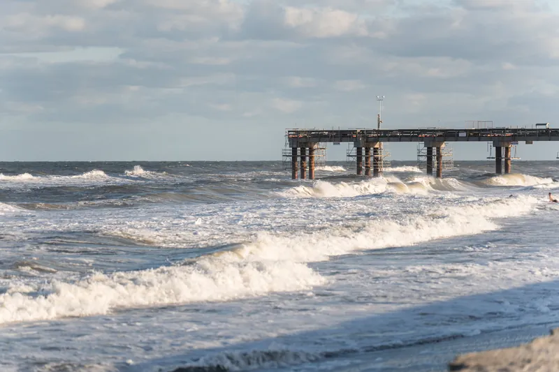 Concrete pier pilings rise from churning surf and foam-topped waves at St. Augustine Beach under overcast sky.