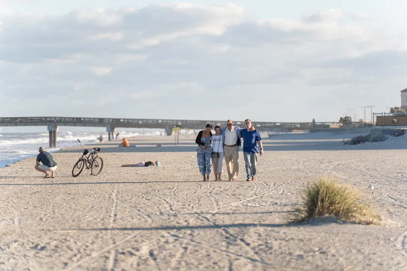 A small group walks across the beach at St. Augustine Beach, with a bicycle nearby and the bridge visible in the distance.