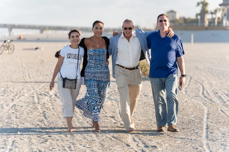 Four people walk arm-in-arm across the sand at St. Augustine Beach with the waterfront and buildings visible behind them.
