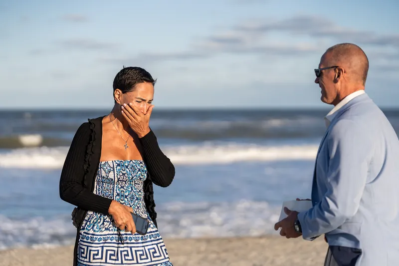 A person in a blue patterned top covers their face with their hand as another person in a light blue shirt stands facing them on the beach.