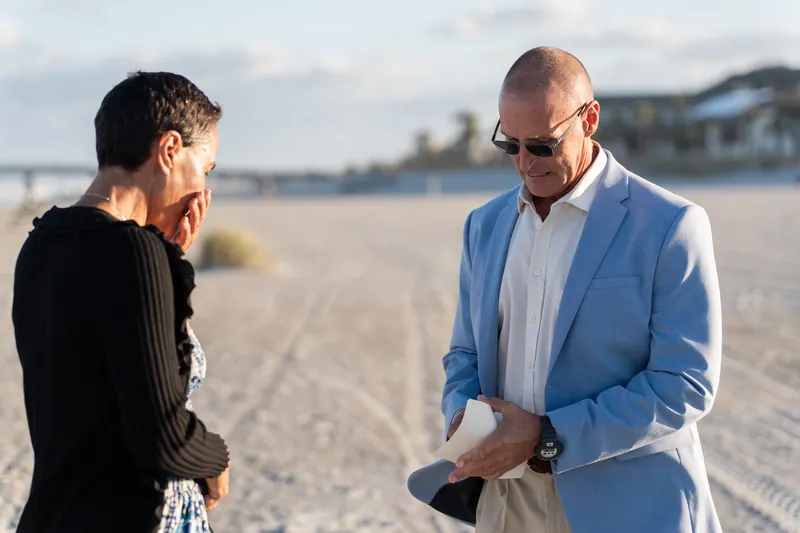 A person in a blue blazer holds an open ring box while facing their partner on the sand.
