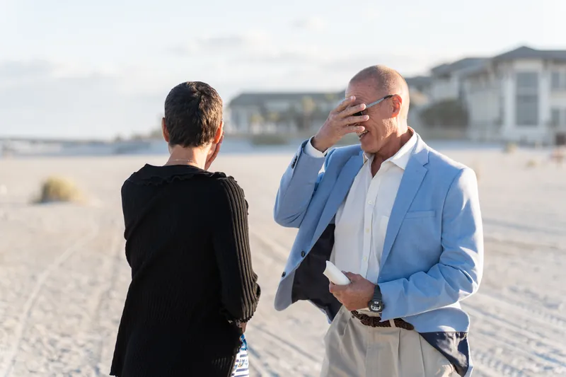 The person proposing extends the ring box as their partner reacts with emotion on the beach.