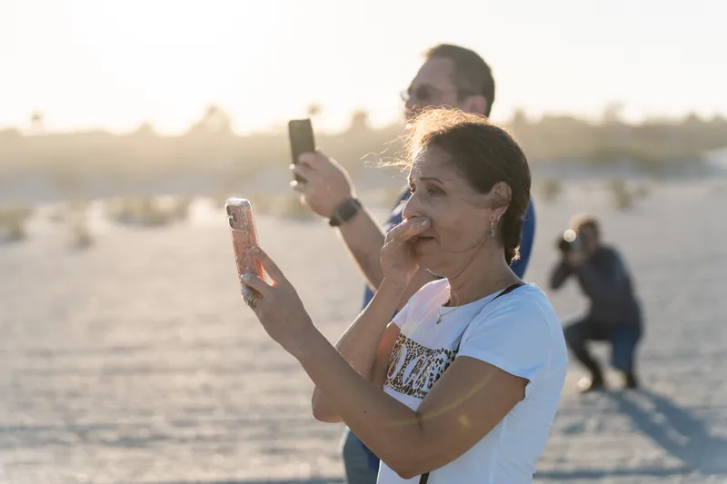 Two people hold up phones at St. Augustine Beach as golden light spreads across the water behind them.