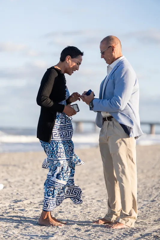 Ella looks down at the ring on her hand while Jeff holds the box, both standing on the sand with breaking waves at St. Augustine Beach.