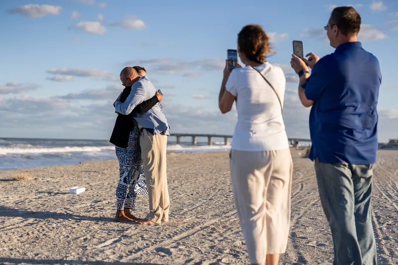 Four people stand on St. Augustine Beach holding phones toward a couple embracing in the sand, ocean and pier visible behind them.