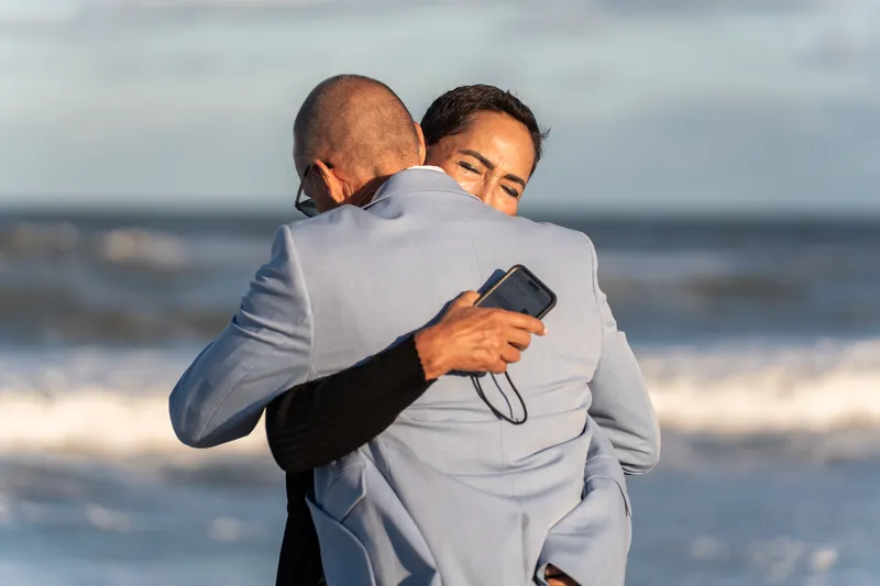 One person hugs another from behind while holding a phone at St. Augustine Beach, waves rolling in the distance.
