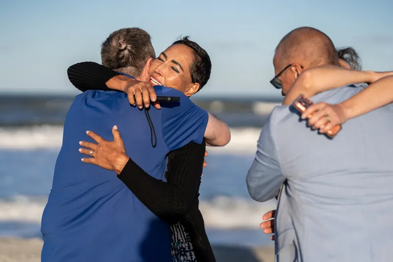 Someone in a blue shirt embraces a person in a patterned dress on the beach as another person reaches in for a group hug.