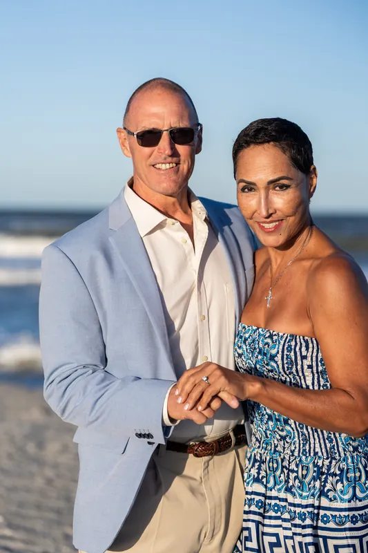 Jeff and Ella hold hands on St. Augustine Beach, both smiling at the camera with the ocean and sandy shore behind them.