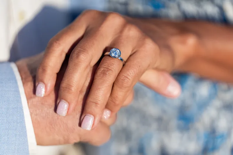 Light blue oval-cut diamond ring on a hand resting against someone's arm at the beach.