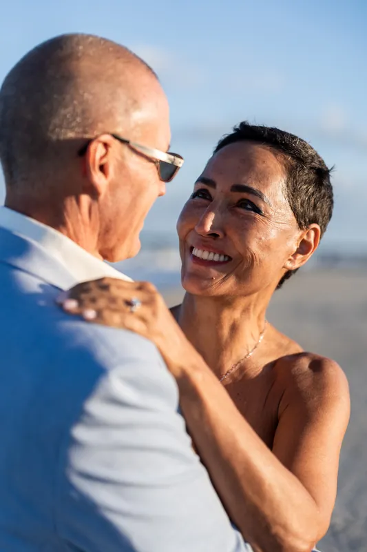 Two people embrace and smile at each other with the ocean and sky behind them.