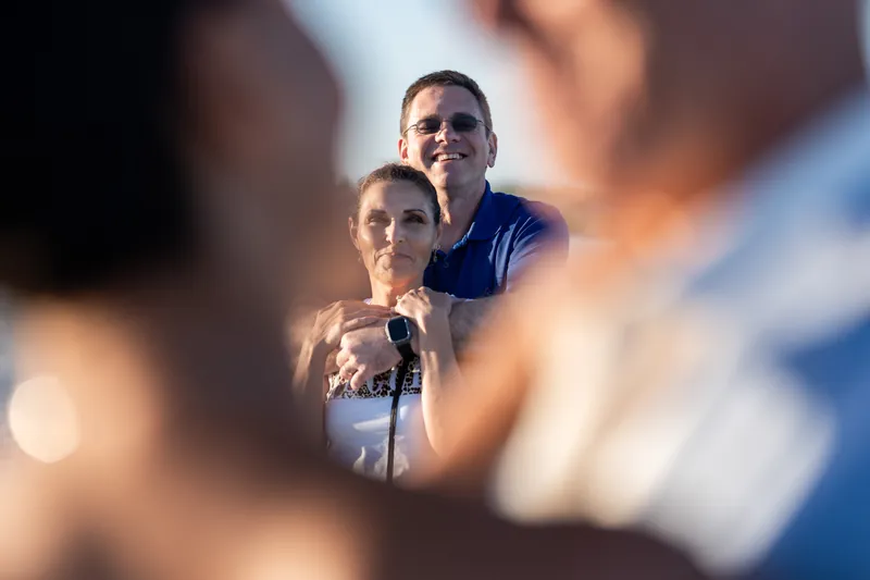 Two people stand together framed by blurred figures in the foreground at the beach.