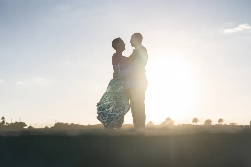 Backlit by sunset, two people embrace while standing in beach dunes.