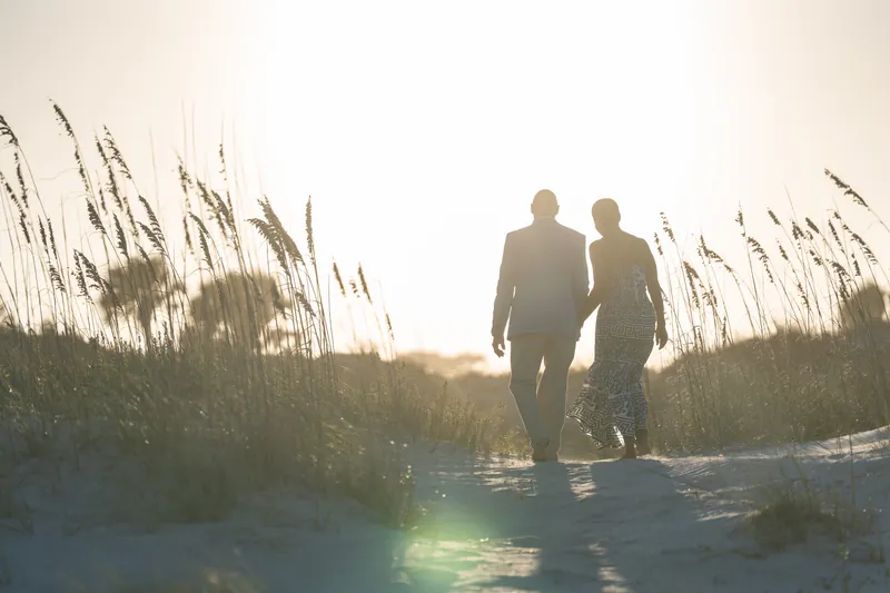 Two figures walk hand-in-hand along a sandy path lined with tall beach grasses at St. Augustine Beach.