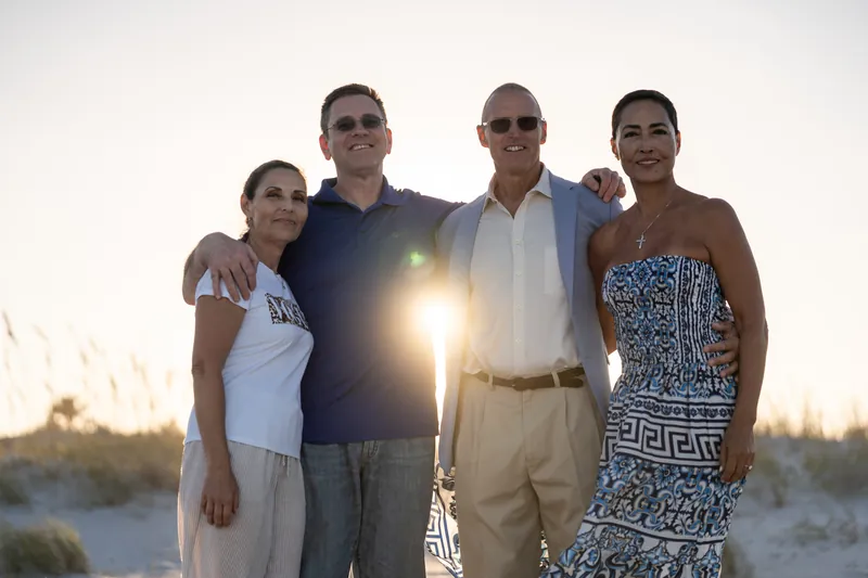 Four people stand together on St. Augustine Beach at golden hour, with the sun flaring between them and ocean dunes behind.