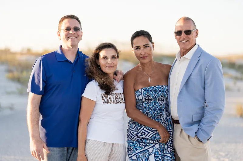 Four adults pose shoulder-to-shoulder on the sand at sunset, smiling at the camera with soft beach light behind them.