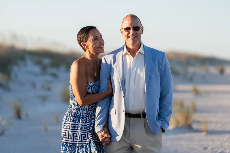A man in a light blue shirt and woman in a patterned dress stand close together on the beach, both wearing sunglasses.