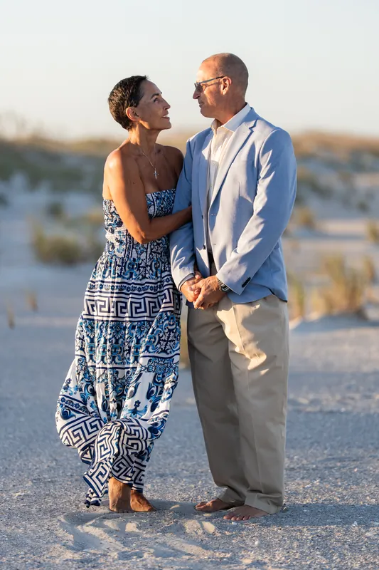The couple holds hands on the sand, facing each other with the beach and dunes glowing in evening light behind them.