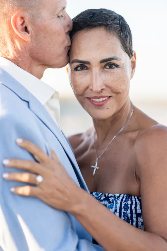 A man in a pale blue shirt kisses a woman's cheek as she smiles, her cross necklace visible against the beach backdrop.