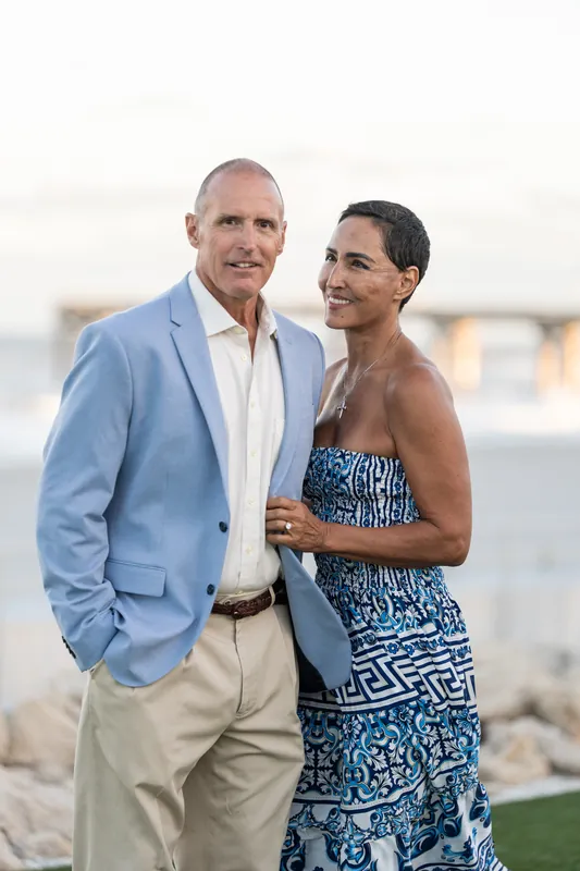 Two people in formal attire embrace on the beach, the pier visible across the water behind them.