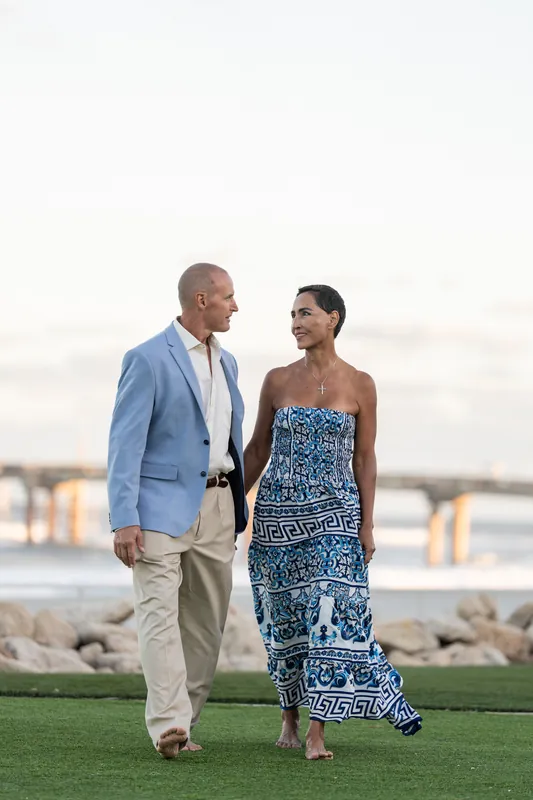Two people walk side by side across a lawn, one in a light blue blazer, the other in a patterned strapless dress.