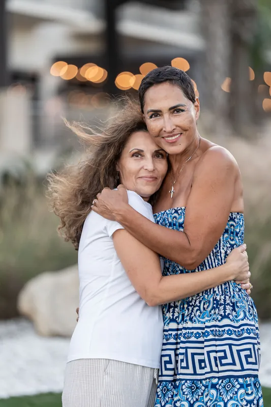 A woman in white hugs the person in the blue patterned dress from behind, both smiling at St. Augustine Beach with string lights blurred above.