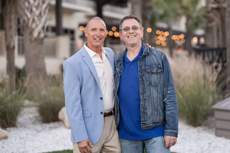 Two men smile at the camera against an evening backdrop at St. Augustine Beach, one in a light blazer and one in a blue shirt with denim jacket.