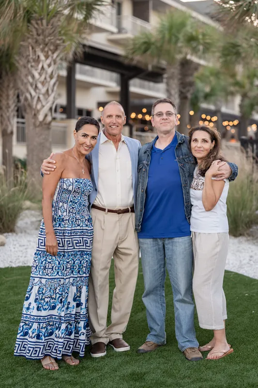 Four adults stand together on grass with palm trees and lit building architecture framing the scene.