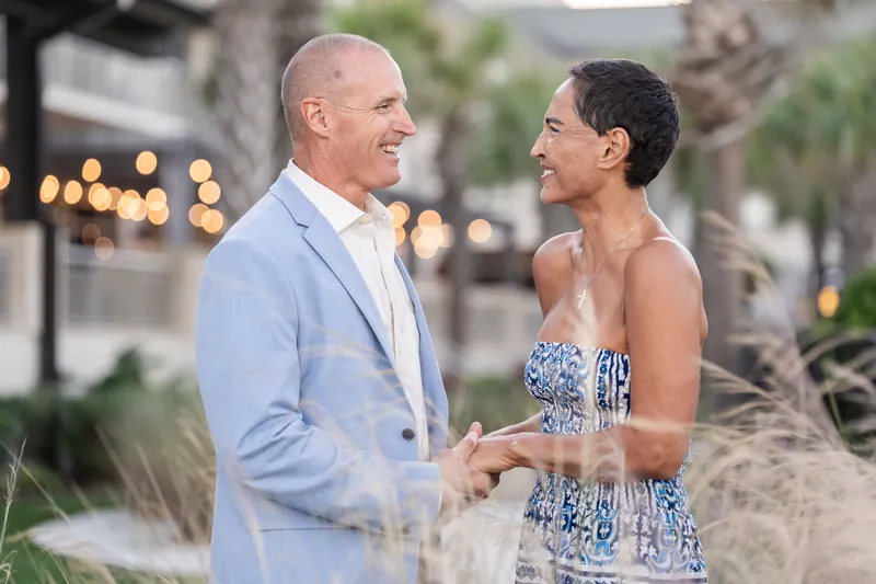 A couple holds hands while facing each other in beachside dunes, palms and colonial buildings softly blurred nearby.