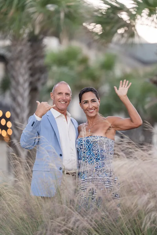 Someone in a light blue suit and patterned dress celebrates with raised hands among beach grasses at dusk.