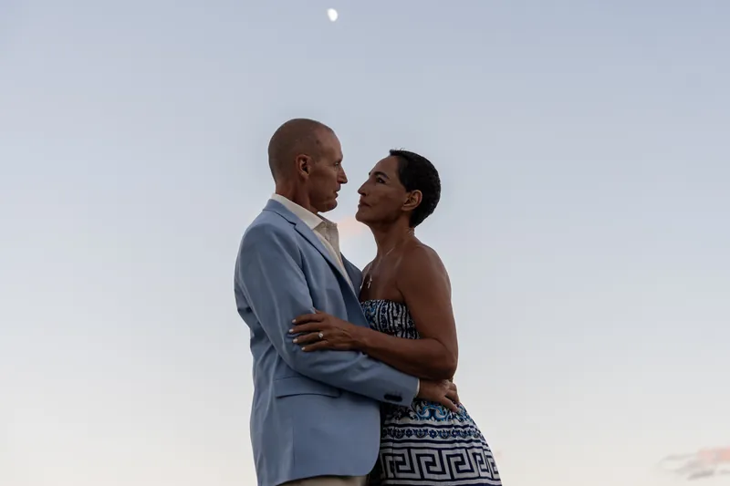 A couple stands close together against an evening sky at the beach, both smiling with the ocean and horizon behind them.
