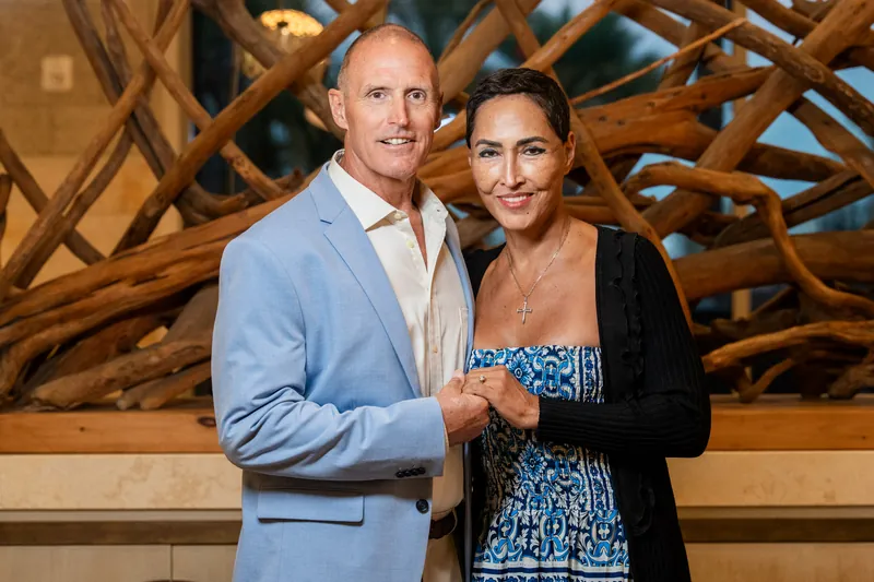 A couple stands arm-in-arm indoors against an ornate wooden architectural backdrop with curved, interlocking design elements.